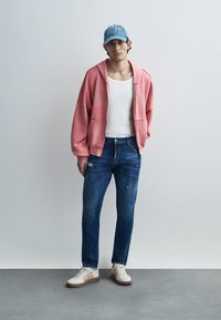 Ροζ φαρδιά μπλούζα με κουκούλα, λευκό αμάνικο τοπ, μπλε τζιν παντελόνι με πιτσιλιές μπογιάς και μπεζ αθλητικά παπούτσια με σχεδιαστές λεπτομέρειες. Μπλε καπέλο.