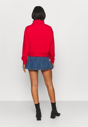 Femme debout de dos, portant un haut rouge à manches longues, une jupe plissée en jean bleu et des bottines noires à talons.