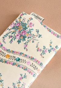 Floral-patterned wallet in cream, featuring colorful flowers, leaves, and decorative borders. Made of textured fabric with a stitched label.