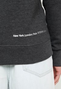 Dunkelgrauer Pullover mit gerippten Bündchen an Ärmeln und Saum. Am unteren Rand befindet sich weißer Schriftzug mit den Worten „New York.London.Paris.“.