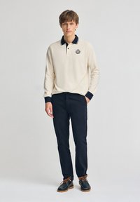 Helles beige Poloshirt mit dunkelblauem Kragen, versehen mit einem Logo. Kombiniert mit dunkelblauen Hosen und dunklen Schuhen, zeigt es einen klassischen, lässigen Look.