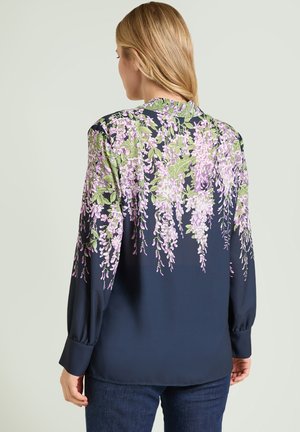 Kvinde iført en marineblå bluse med faldende lilla og grønne blomsterprint, stående med ryggen mod en neutral baggrund.