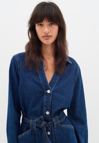 Denim haljina košulja u tamnoplavoj boji s dugim rukavima, prednjim kopčanjem na gumbe i vezanjem u struku. Sadrži dvije prednje džepove.