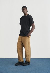 Μαύρο κοντομάνικο μπλουζάκι, καφέ φαρδιά παντελόνια και μαύρα αθλητικά παπούτσια. Το μπλουζάκι έχει λογότυπο στο γιακά. Ανθεκτικά υλικά με casual σχεδιασμό.