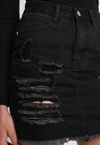 Černá džínová sukně s roztrhanými otvory, třepenými okraji a přiléhavým střihem, doplněná předními kapsami a knoflíkovým zapínáním.