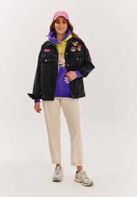 Czarna dżinsowa kurtka z naszywkami, fioletowy hoodie z rysunkiem kreskówkowym, beżowe spodnie i szare trampki, dopełnione różową czapką.