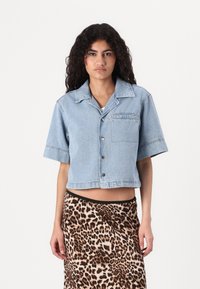 Jaqueta de ganga azul cropped com mangas curtas, fecho com botões e um bolso no peito, combinada com uma saia de padrão de leopardo. Textura suave.