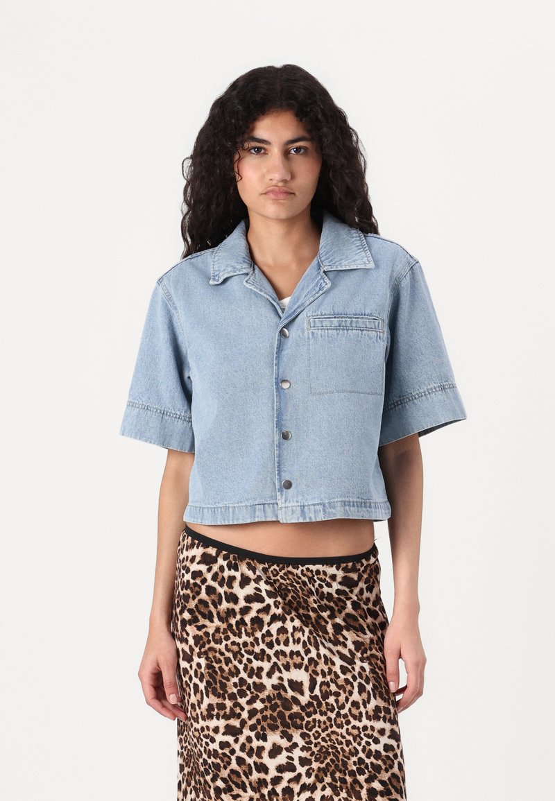 Jaqueta de ganga azul cropped com mangas curtas, fecho com botões e um bolso no peito, combinada com uma saia de padrão de leopardo. Textura suave.