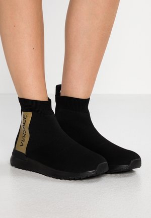 Baskets de style chaussette noires avec des bords côtes à la cheville et des panneaux verticaux dorés du logo VERSACE portées sur des jambes nues sur un fond uni.