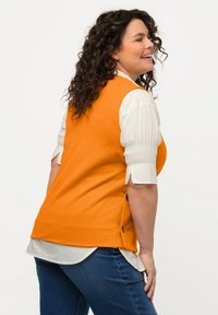 Oranje mouwloos vest met zijknopen, gedragen over een witte blouse met pofmouwen, gecombineerd met blauwe jeans. Glad textuur en getailleerd ontwerp.