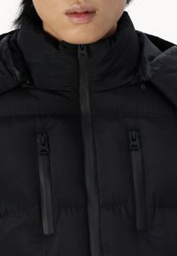 Schwarze Pufferjacke mit hohem Kragen, ausgestattet mit drei Reißverschlusstaschen und glatter Textur. Die Reißverschlüsse sind alle schwarz und ohne sichtbare Logos.