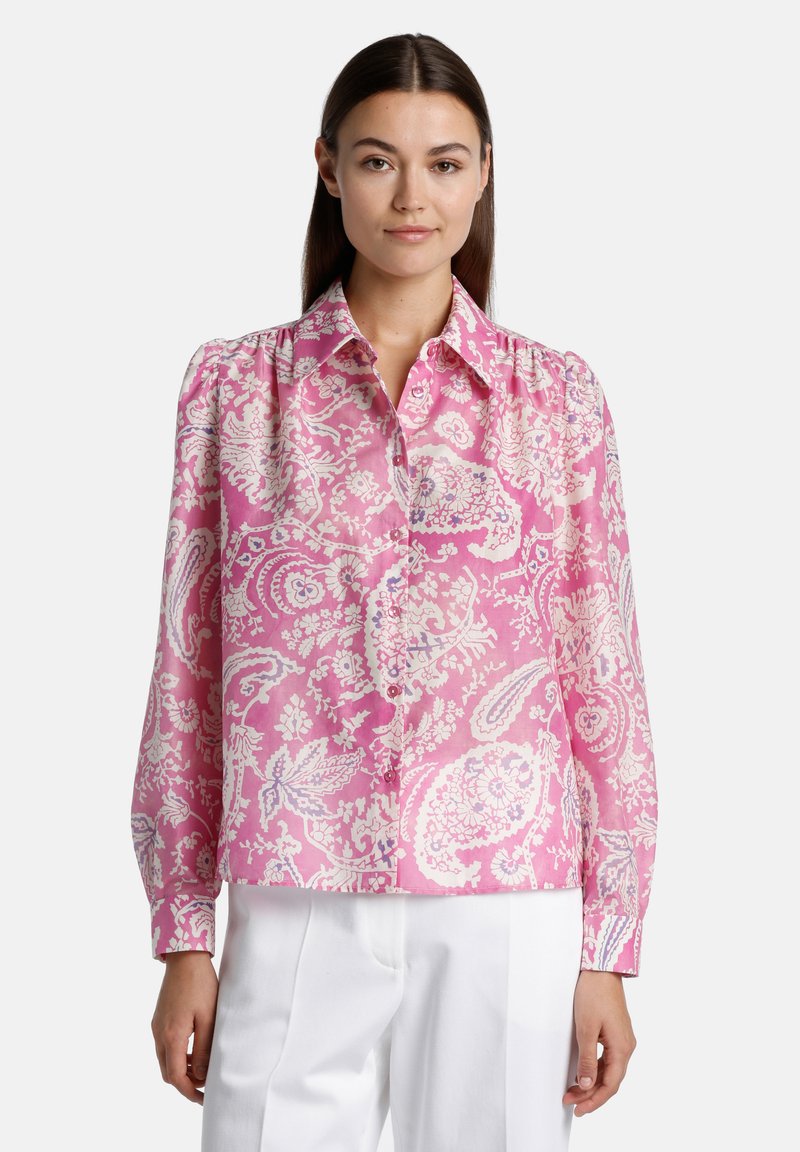 Blusa rosa a fantasia con un motivo paisley, chiusura con bottoni frontale, colletto a punta e maniche lunghe, realizzata in un tessuto liscio. Abbinata a pantaloni bianchi.
