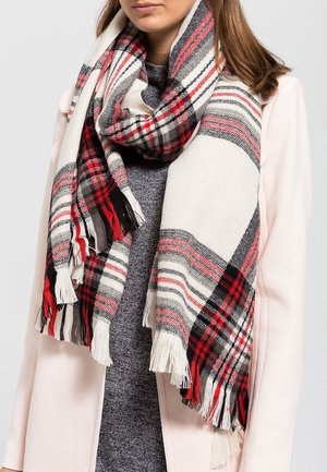 Femme portant une écharpe à carreaux rouge, noir et blanc avec des franges, par-dessus un haut gris et un manteau rose clair.