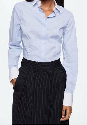 Mujer con una camisa azul celeste abotonada con puños blancos y pantalones negros de rayas de talle alto, manos parcialmente en los bolsillos.