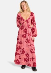 Vrouw in een roze maxi-jurk met lange mouwen, versierd met rode bloemenpatronen en uitsparingen, staand met één hand op haar heup, met zwarte schoenen.