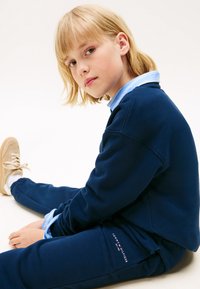 Marineblauwe sweatshirt- en broekenset van zachte stof, voorzien van een lichtblauw overhemd met kraag eronder en logodetail op de broek.