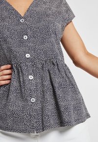 Kvinde iført en kortærmet, marineblå mønstret bluse med hvide knapper og hvide bukser, hånd hvilende på hoften.