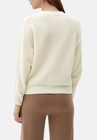 Cremefarvet uldtrøje med en tekstureret strik, brede ærmer og ribbet kant. Har en rund hals og slutter ved taljen, båret sammen med brune bukser.