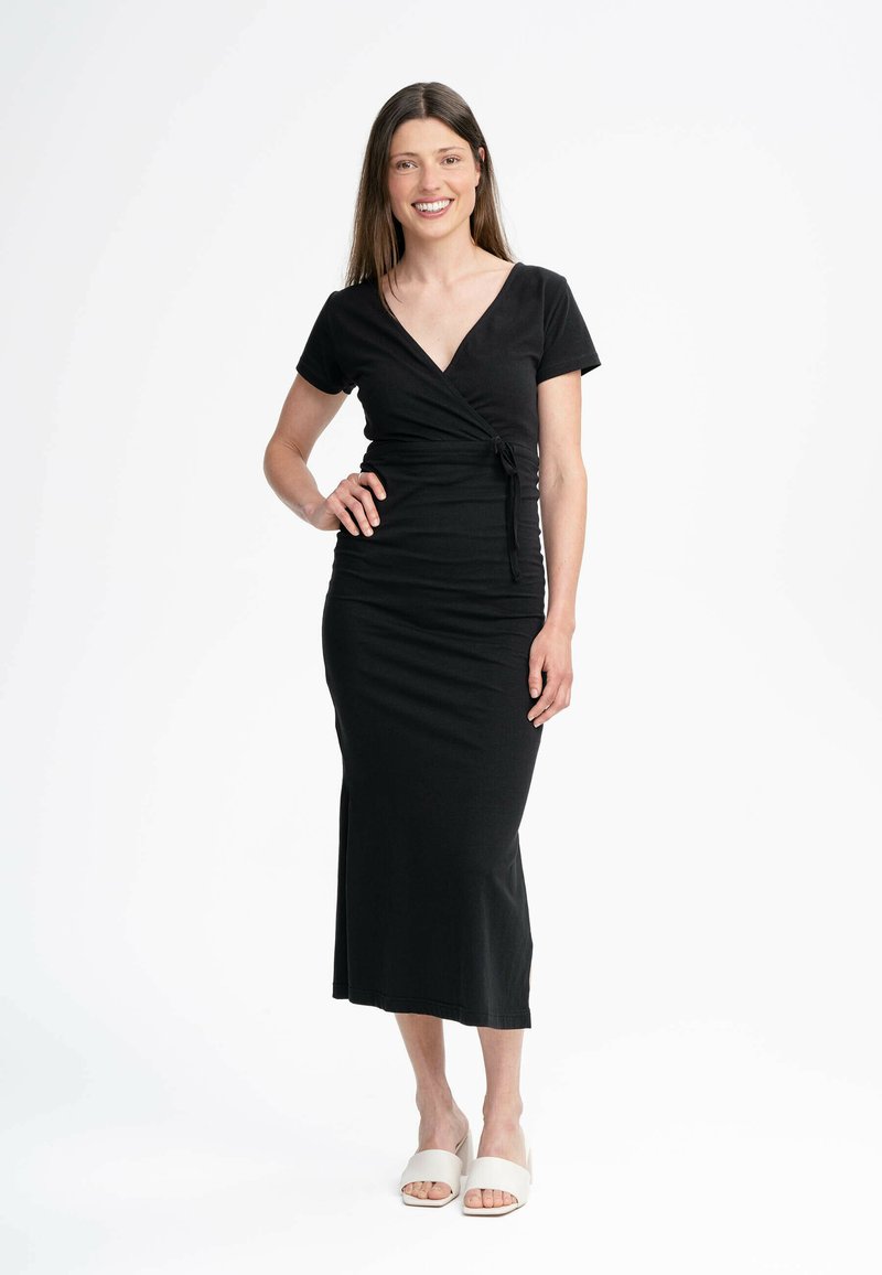 Schwarzes Wickelkleid mit kurzen Ärmeln, V-Ausschnitt, Raffung in der Taille und einer figurnahen Silhouette, die bis zur Mitte der Wade reicht. Getragen mit beigen Blockabsatzsandalen.