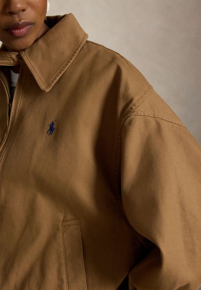 Primo piano di una persona che indossa una giacca marrone con un logo ricamato di un giocatore di polo blu sul petto a sinistra e un ampio colletto.