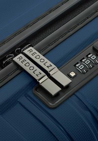 Primo piano delle cerniere di una valigia blu etichettate "REDOLZ" accanto a un lucchetto nero a combinazione impostato su 0-0-0.