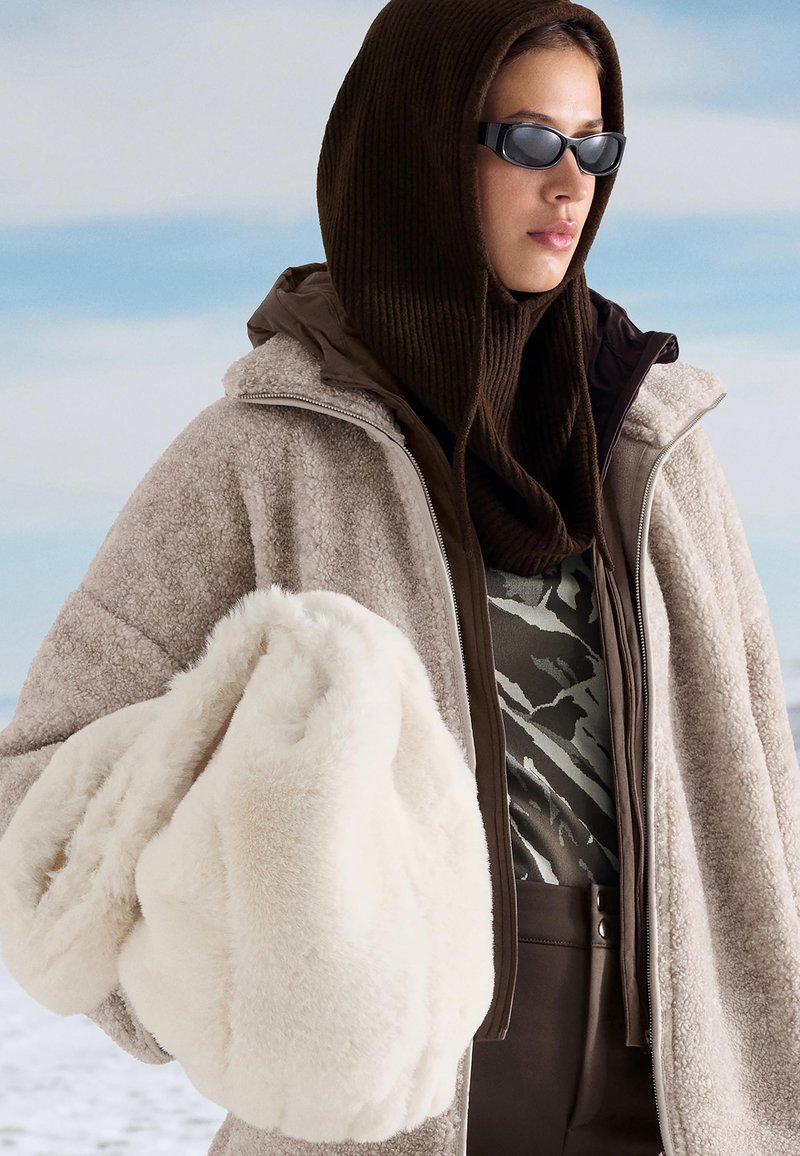 Žena nosi bež flis jaknu, smešnu maramu s kapuljačom, sunčane naočale i drži veliku belu pahuljastu torbu u snežnoj vanjskoj sredini.