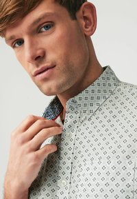 Camisa abotonada con una tela de color claro que presenta un patrón geométrico en azul marino. Cuello de contraste con un patrón visible y detalles de botones.