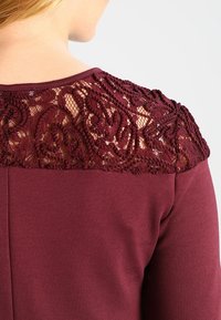 Primer plano de la parte trasera del hombro de una blusa burdeos con un intrincado encaje floral superpuesto y una tela sólida debajo.