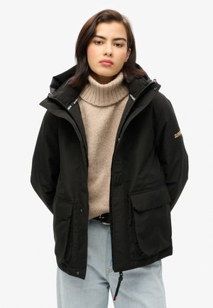 Zwarte waterdichte jas met capuchon, ritssluiting en twee voorzakken, gedragen over een lichtbeige gebreide trui. Kenmerkt logo-accenten.