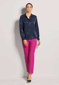 Bluse mit maritimem Muster und langen Ärmeln, mit Knopfkrause, kombiniert mit figurbetonten pinken Hosen und schwarzen High Heels. Glattes Material, maßgeschneiderte Passform.