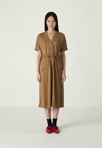 Rochie maro, până la genunchi, cu mâneci scurte, decolteu în V, închidere cu nasturi, două buzunare frontale și curea legată la talie. Asortată cu pantofi roșii și șosete negre.