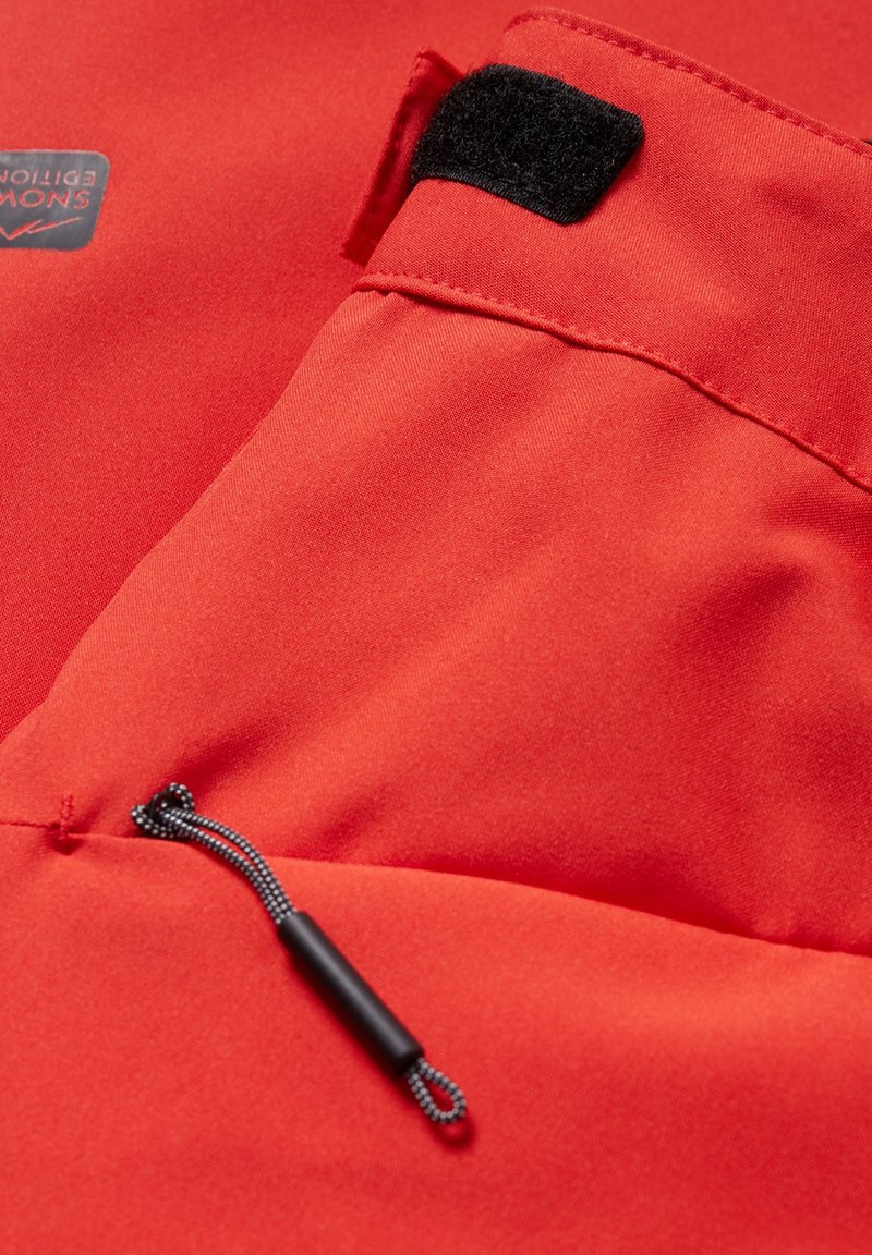 Roter Stoff mit glatter Textur, versehen mit einem Klettverschluss, Taschendetails und einem schwarzen Zugband mit einem Stopper zur Anpassung.