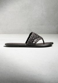 Sandale plate noire avec bride en cuir tressé et entre-doigt, vue de côté sur une surface et un fond gris.