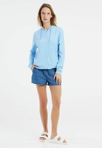 Felpa azzurra chiara con zip, tessuto morbido, con una tasca frontale. Abbinata a pantaloni shorts in denim e sandali bianchi. Design semplice e casual.