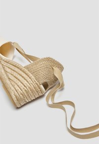 Sandale compensée en toile beige avec une plateforme en jute tissé, des brides en tissu et une semelle plate. Texture lisse avec des détails tressés sur la plateforme.