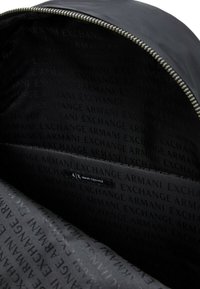 Interior de mochila de tela negra con una textura suave. Cuenta con un compartimento grande y un bolsillo lateral, forrado con un patrón repetido del logo.