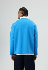 Blauwe sweatshirt met een witte kraag, voorzien van lange mouwen en een losse pasvorm. De stof lijkt zacht; gedragen over een donkere spijkerbroek.