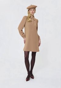 Frau in einem camel-farbenen Kleid mit Puffärmeln und Knöpfen, passendem Barett, schwarzen Strumpfhosen und burgunderfarbenen Absätzen, stehend vor einem schlichten Hintergrund.