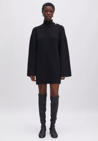 Schwarzes Strickkleid mit hohem Kragen, oversized Ärmeln und goldenen Knopfakzenten an der Schulter. Kombiniert mit kniehohen schwarzen Stiefeln.
