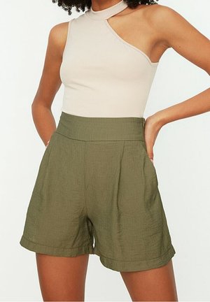 Femme portant un top beige à une épaule et un short taille haute vert olive avec poches latérales, se tenant les mains sur les hanches.