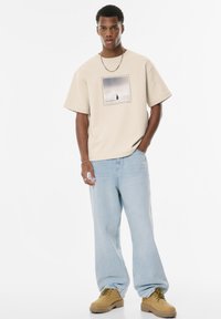Beige oversized t-shirt med et sort-hvidt grafik på forsiden, parret med lyseblå straight-leg jeans og brune arbejdsstøvler.