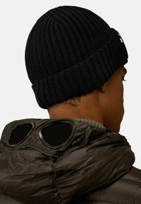 Berretto di lana nera con un design a coste, indossato sulla testa. Sullo sfondo si vede un giubbotto imbottito marrone con accenti neri simili a occhiali.