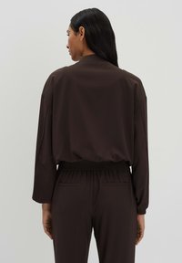 Brown lightweight jacket with a relaxed fit, long sleeves, and elastic waistband, shown from the back. Smooth texture, minimalistic design.