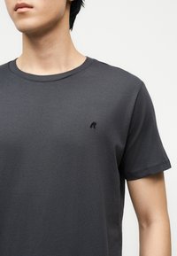 Νεαρό άτομο που φοράει απλό σκούρο γκρι T-shirt με στρογγυλή λαιμόκοψη και μικρό μαύρο λογότυπο "R" στο αριστερό στήθος, πάνω σε λευκό φόντο.