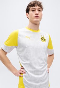 Λευκή και κίτρινη ποδοσφαιρική φανέλα Puma με στρογγυλή λαιμόκοψη, κοντά μανίκια, υφασμένο σχέδιο και λογότυπο της ομάδας στο στήθος.