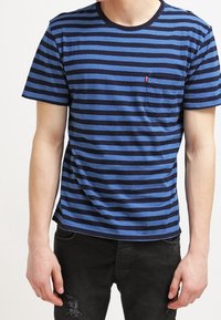 Blå och marinrandig t-shirt i bomull; har rundad hals och en liten bröstficka på vänster sida. Matchas med svarta jeans.