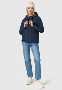 Marineblå pufferjakke med hætte, der har lynlås, elastiske manchetter og to forneden. Bæres over lyseblå jeans og sneakers.