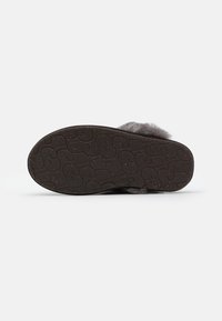 Semelle d'une pantoufle foncée et duveteuse avec un dessous en caoutchouc texturé montrant un motif répétitif pour l'adhérence sur fond blanc.