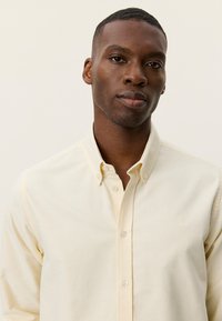 Ανοιχτό κίτρινο πουκάμισο με γιακά και κουμπιά, κατασκευασμένο από λείο ύφασμα· διαθέτει διακριτικό κεντητό λογότυπο στο στήθος και κούμπωμα με κουμπιά.