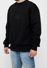 Sudadera negra con cuello redondo, puños y dobladillo acanalados, que presenta un sutil diseño de texto impreso en un tono oscuro. Lleva puesta con jeans de color azul claro.